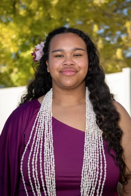 Kahealani Blackmon wears a purple dress and white beaded necklace, smiling at the camera. 