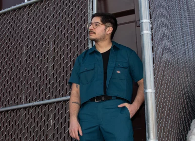 A bespectacled person with a mustache stands in front of a brown cement building encased by a chainlink fence. They wear a a blue worksuit combo from Dickies.