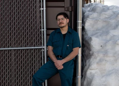 A bespectacled person with a mustache stands in front of a brown cement building encased by a chainlink fence. They wear a a blue worksuit combo from Dickies.