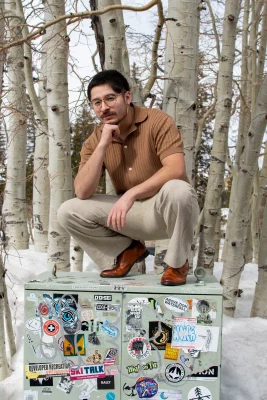 A bespectacled person in khaki colored pants and brown leather loafers squats atop a sticker covered powerbox. Trees lining behind them.