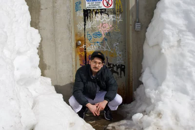 A bespectacled person with a mustache stands in front of a graffitied cement door surrounded by snow. They wear an outfit reminiscent of a Baseball player's uniform.