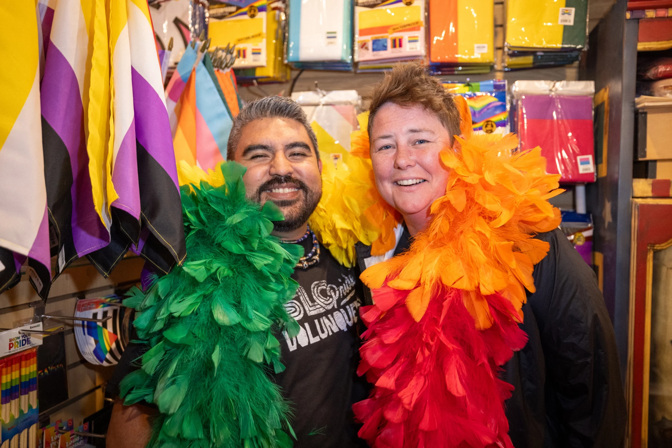 Roberto Lopez and Bonnie O’Brien smile into the camera, they wear colorful feather boas.