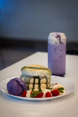 A stack of Japanese fluffy pancakes surrounded by strawberries and ice cream. Behind it, an Ube latte in a glass.