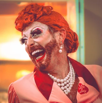 A drag queen imitating Lucille Ball laughs off to the side of the camera. She has a red glittery beard.