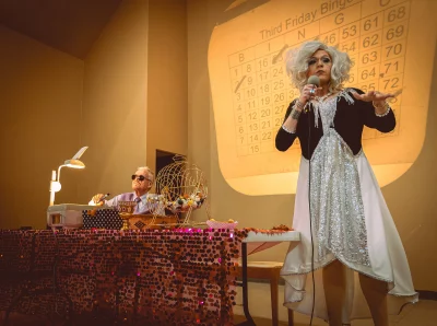 A drag queen in a giant white wig and long flowey dress stands in front of a bingo caller, she holds a microphone.