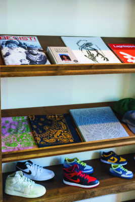 Three shelves along a white wall containing books, magazines, bandanas and sneakers, respectively. 