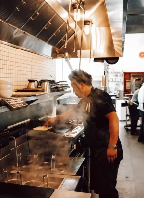 A man in a black uniform stands in a kitchen. He flips a portion of a dish on a grill.