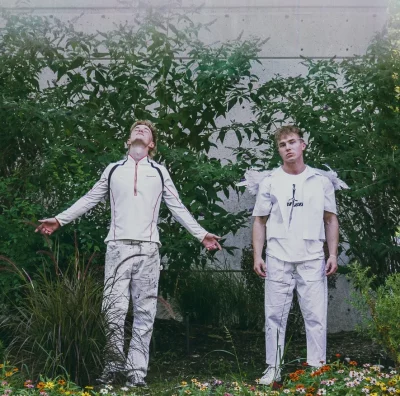 Two blonde men, dressed in all white, pose in front of foliage. 