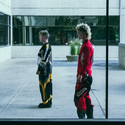 Two blonde men stand and look off to the left. They're dressed in motocross uniforms.