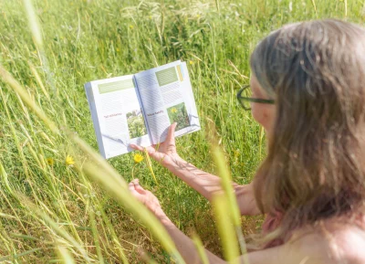Lisa Thompson comparing a dandelion while crouched in a filed of grass to her photos of dandelions inside her book.
