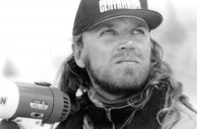 A young Renny Harlin with long hair holds a megaphone and looks off to the side of the camera. He wears a baseball cap with the film logo "Cliffhanger" on it.