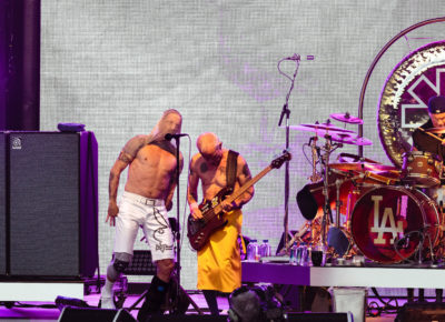Singer Anthony Kiedis lifts his mesh shirt over his head while singing at the mic next to bassist Flea and drummer Chad Smith as they play on stage. Photo: @lmsorenson
