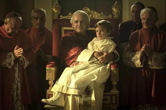 A young boy in white Catholic dressing sits on the lap of a priest. He is surrounded by other clergyman.