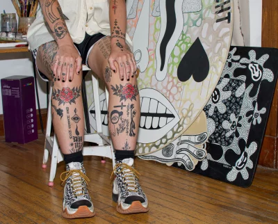 Cake's tattoos on their shins and hands with their artwork in the background. 