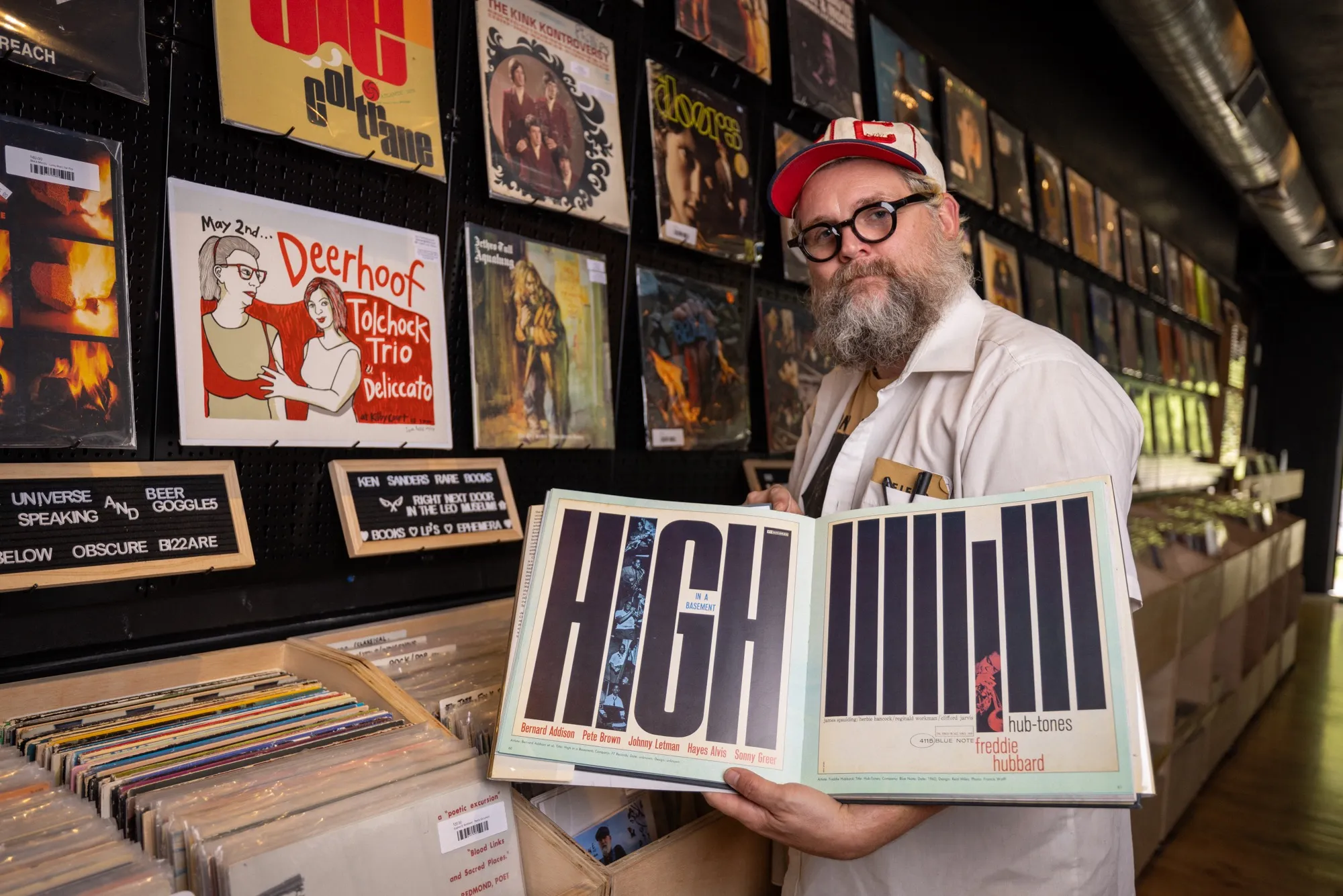 Adam Michael Terry with a red cap, round glasses and beard shows off one of his favorite records in his shop.