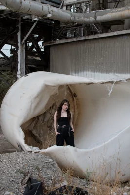 Ophelia May, Mercy Seat, stands to the front of the camera with her hands in her pockets. She stands in what looks to be a half melted industrial plastic bin.