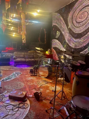 A dim whimsical looking venue space, with lights hanging from the ceiling above a couch in the background and array of musical instruments littered in the foreground. 