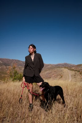 Chloe Akira wears a black jacket and black skirt stands in a field with her black dog.