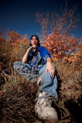 Chloe Akira wears a blue button down dragon shirt and baggy jeans while sitting on grass. 