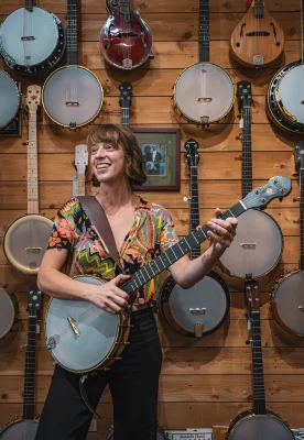 Melissa Chilinski holding a banjo, stands in front of a wood panel wall with banjos and a painting hung on it.