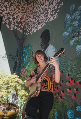 Melissa Chilinski stands in front of a mural with colorful flowers, holding her banjo. 