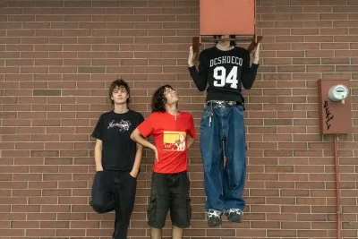 The member of eardrums on the left leans on the brick wall with their hands in their pockets while the one in the middle looks up at their bandmate dangling from a fire escape.