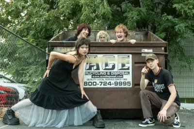 Three members of Lovelace hide in a dumpster while the other two pose in front of them. 