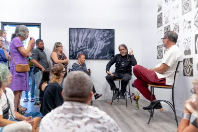 A group of people listen to Russel Albert Daniels as he is talking in front of one of his photographs and a wall of his drawings. 