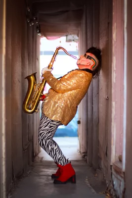 Genda is wearing a sparkly gold jacket and zebra print pants as he plays the saxophone.