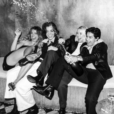 The Aces in a black and white image of the quartet popping a bottle of none labeled champagne. The sit on a tufted bench, jumping and laughing at the popping bottle.