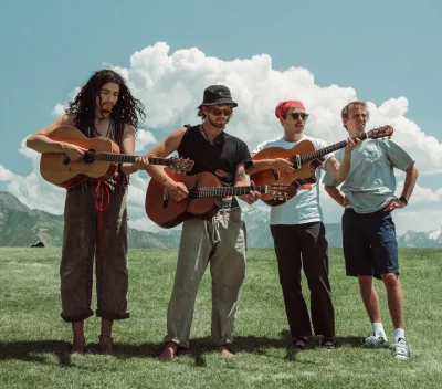 The band The Moss stand in a picture-esque field. Three of the four members hold plain wooden guitars. 