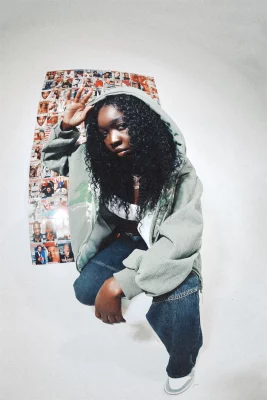 Person wearing hoodie crouching in front of a photo wall in a white room.