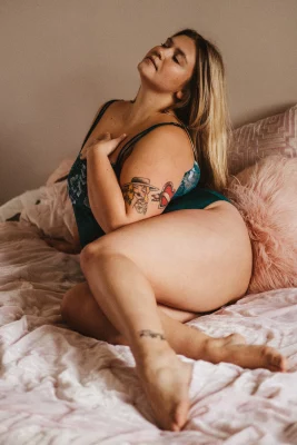 Colored image of a woman with long blonde hair in blue lingerie. She sits on a bed of pink sheets.