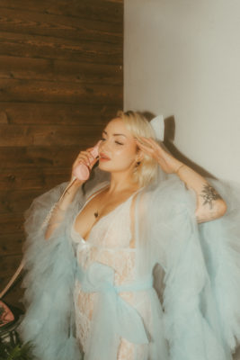 A blonde, white woman talks on a pink wired phone while wearing a blue mesh robe.