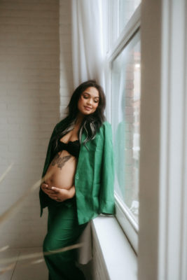 A pregnant woman with a butterfly stomach tattoo leans against a window.