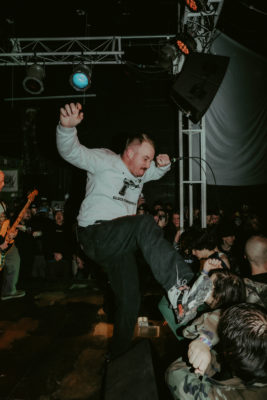 A vocalist on stage in a grey pullover kicks his foot at the audience.