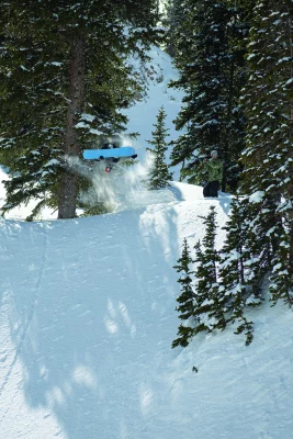 Two snowboarders in the mountains, one is filming while the other is taking a jump off of the hill.