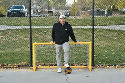 Travis Winn stands in front of a yellow soccer goal post and holds a soccer ball under his foot.