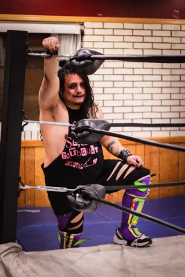 Princess Deathwish hangs off the side of the ring with one leg up and a smile on his face.