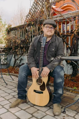 John Elwood Morgan sits and smiles in front of Greenhouse Effect with his guitar in front of him. 