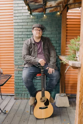 John Elwood Morgan sits on a stool in front of a green brick wall. 